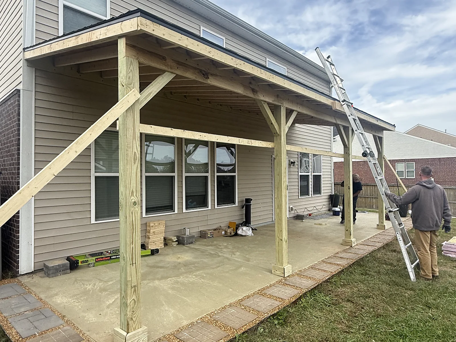 A covered patio under construction, with the timber frame up, a ladder against the roofline, and two crew members working on site.