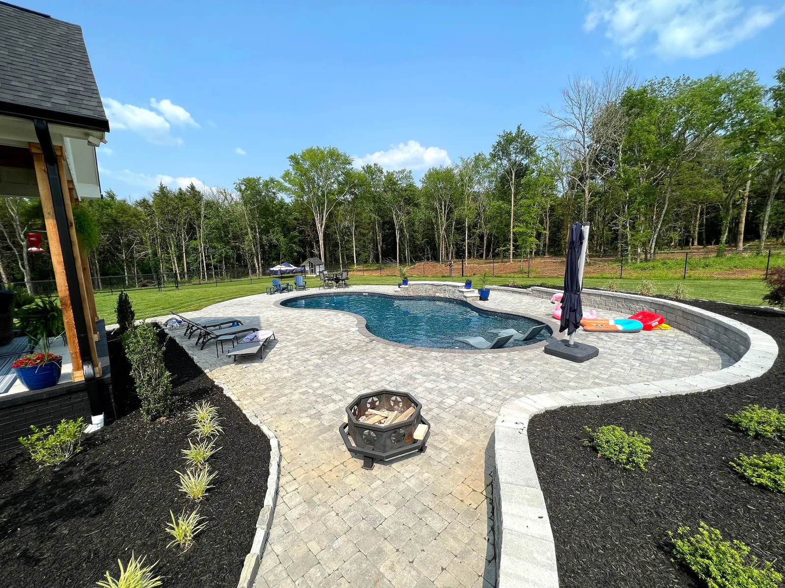 A finished freeform backyard pool with a paver deck, retaining wall, and fire pit, backing onto a Middle Tennessee treeline.